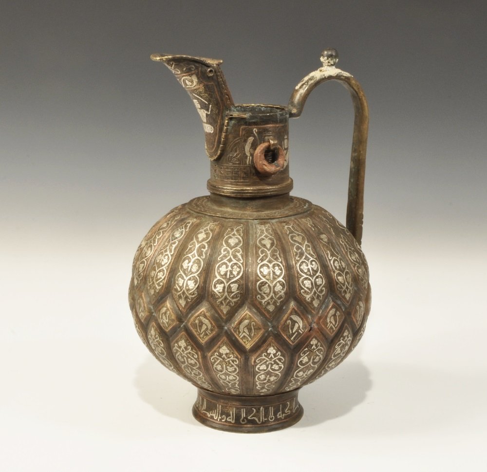 Islamic Bronze Figural Ewer: 19th century AD. A cast ewer with segmented globular body, flared foot, tubular neck with lipped spout and lateral suspension rings, D-section handle to the rear with bulb thumb-pad; silver-inlaid sph