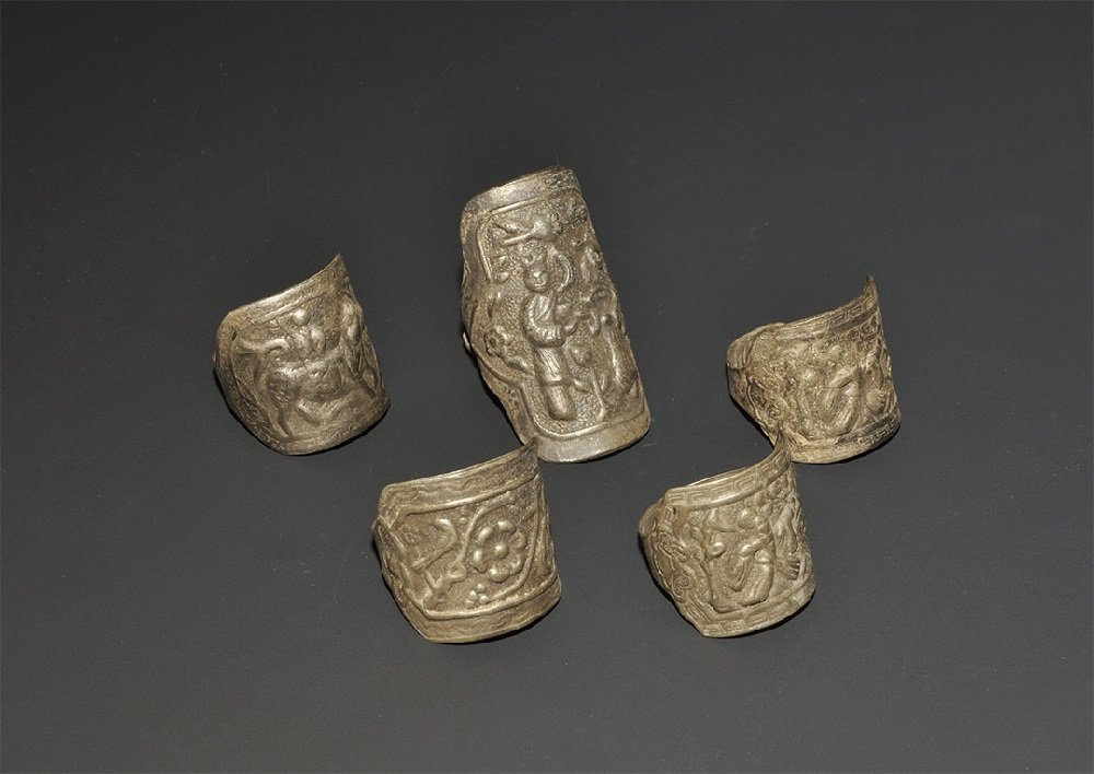 Chinese Silver Coloured Metal Repoussé Ring Group: 20th century AD. A mixed group of finger rings, each with narrow band and repoussé plaque with figural scene. 24 grams total, 22-40mm (3/4 - 1 1/2"). From a central London collection formed between 1