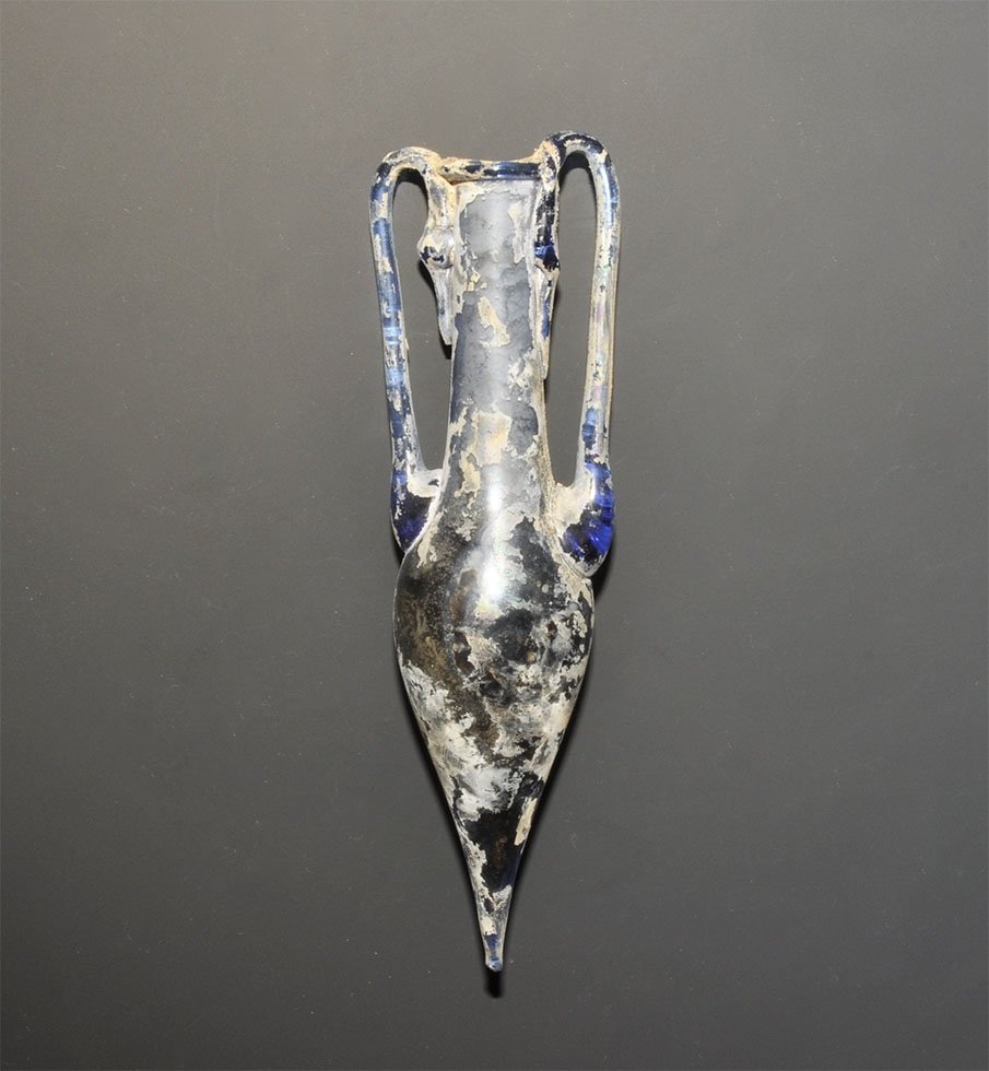 Roman Style Glass Amphora Vessel: An undated archaistic miniature amphora in deep blue glass with some iridescence to the surface; applied trail rim and handles. 56 grams, 15.5cm (6"). Acquired on the London art market in the late 198