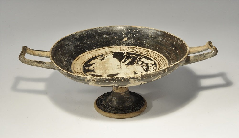 Greek Apulian Painted Tazza: 5th-3rd century BC. A broad buffware dish with pedestal foot and two square handles; black inner face with reserved white figure of a horseman with a lyre within a meander border; to the outer face, p