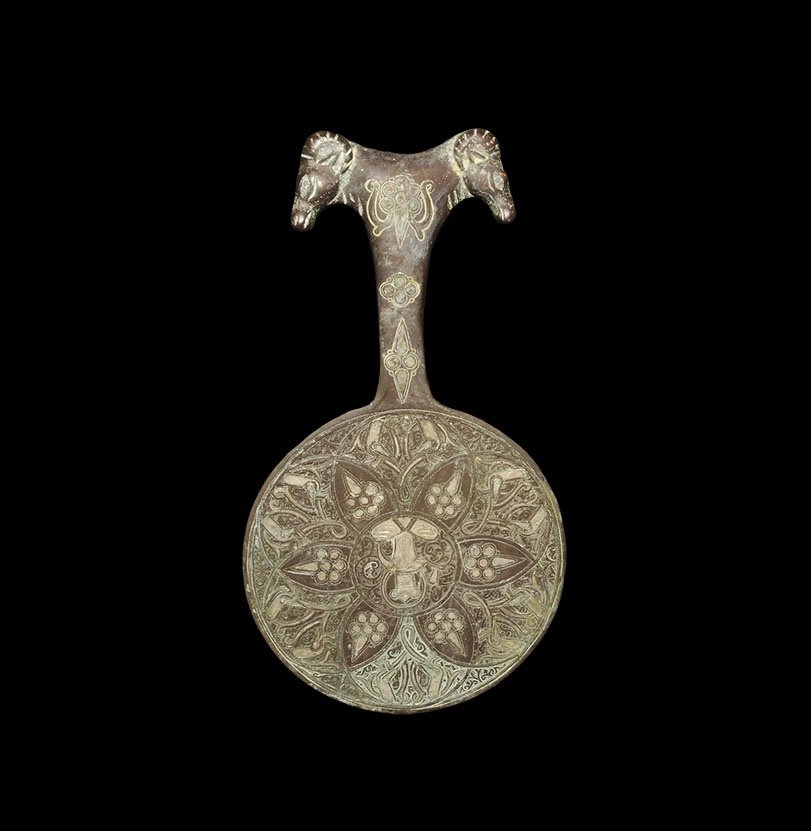 Islamic Inlaid Bronze Handled Mirror: An undated archaistic cast discoid mirror with T-shaped handle, ram's head finials; to the reverse a silver-inlaid seated figure with a cup surrounded by a band of arabesque ornament. 498 grams, 22cm