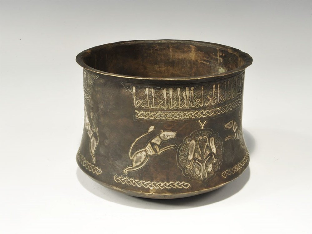 Islamic Gilt-Bronze Bowl with Silver Calligraphic Inlay: An undated archaistic carinated bowl with flange rim; silver-inlaid roundels, each with birds and leaping hounds, guilloche above and below, panel of calligraphic text. 714 grams, 15cm (5 3/4"). Prope