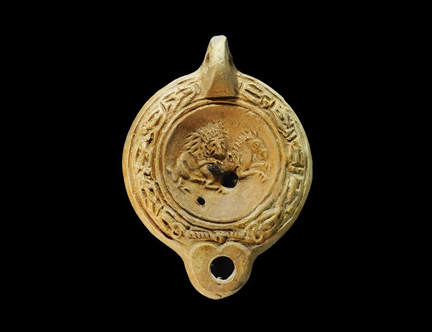 Roman Ceramic Lion and Horse Oil Lamp: 1st century AD. A squat oil lamp with heart-shaped nozzle, pierced lug handle to the rear; band of laurel leaves to the border enclosing a scene with a lion attacking a horse; rosette to the underside