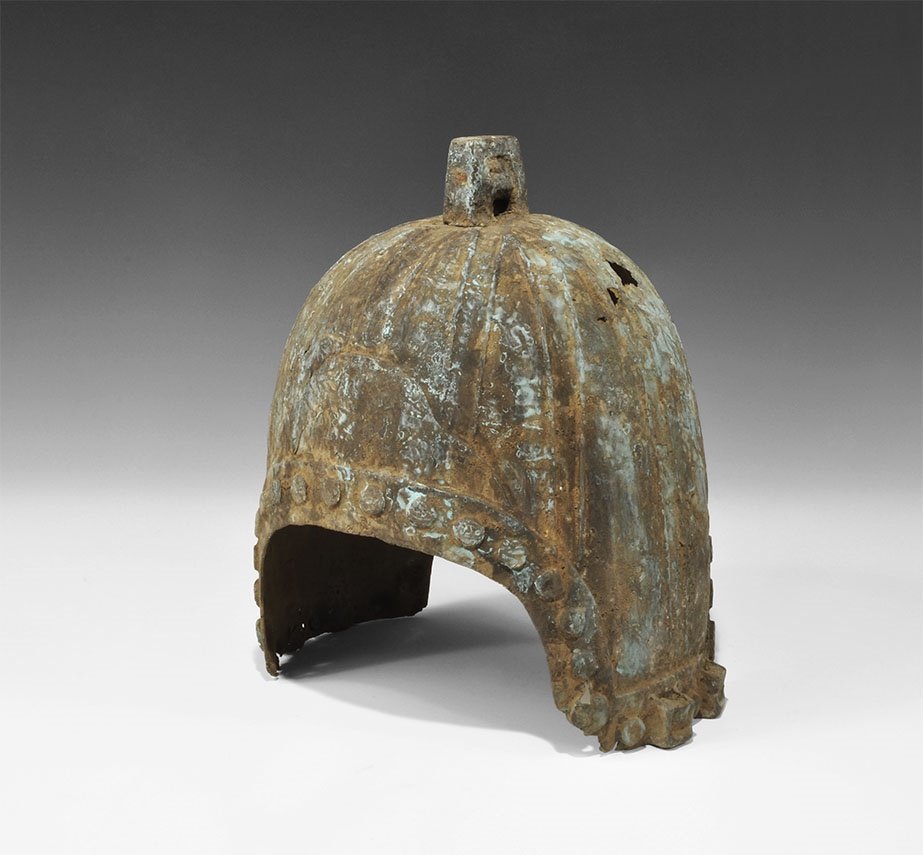 Chinese Bronze Zhou Dynasty Helmet: 800-500 BC. A cast bronze helmet of unusual design, with D-shaped openings to front and rear; the bowl formed with longitudinal ribbing, reinforcing plates and simulated rivet detailing; above, a pier