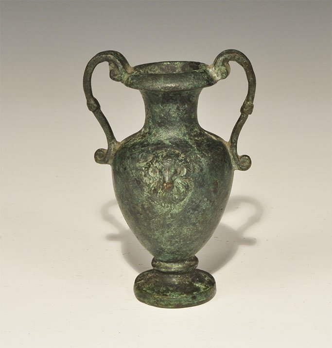 Greek Bronze Vessel with Ram's Heads: An undated archaistic cast bronze vase with flared base, chamfered rim and two scrolled handles; to each face a ram's head mask. 357 grams, 11 cm (4 1/4"). Property of a Cambridgeshire gentleman. Time