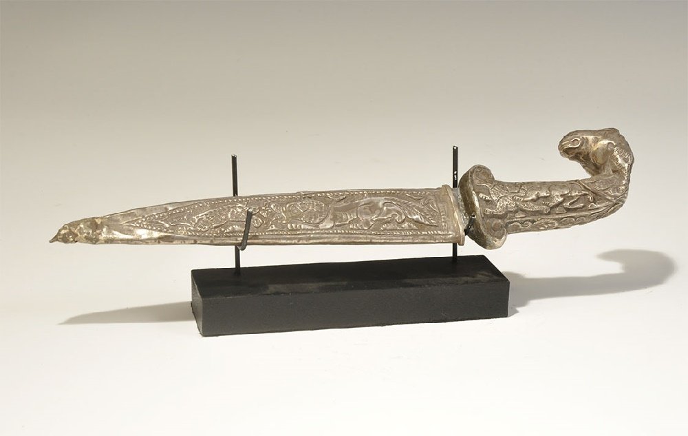 Indian Dagger with Silver Ram's Head Hilt: 20th century AD. A dagger and scabbard comprising a triangular blade with incised zoomorphic decoration to each face, grip with scenes of animal predation, curved pommel formed as a ram's head; scabba
