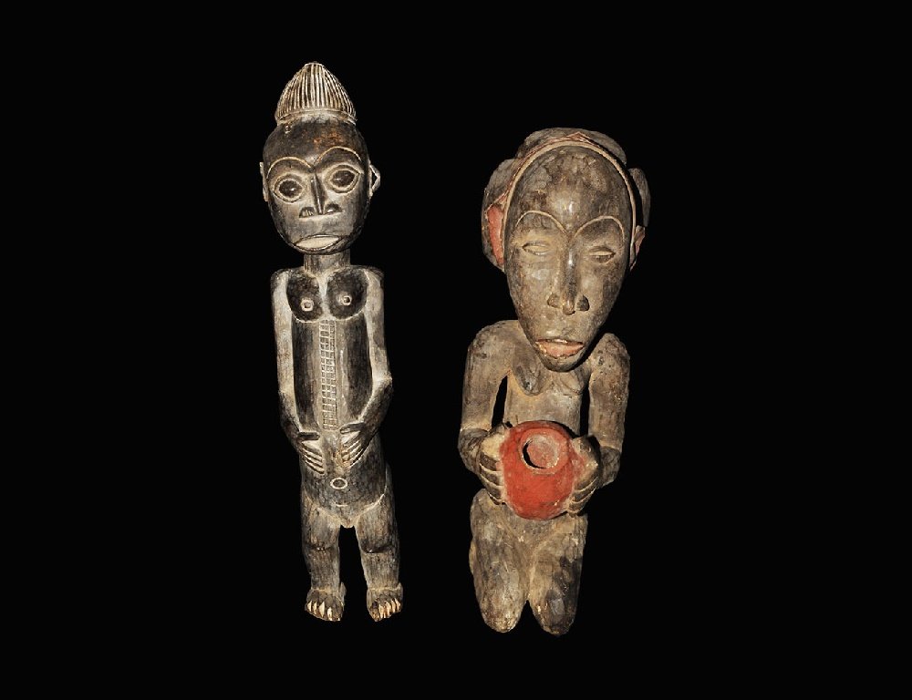 Ethnographic Wooden Statue Pair: 20th century AD. A group of two tribal figures comprising: an Ivory Coast (Baule) carved standing figure of a pregnant woman with stylised facial features, vertical band of scarification (?) to the ab