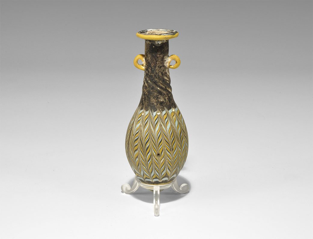 Roman Style Mosaic Glass Vessel: 20th century AD. A piriform unguentarium with rolled rim and two lateral applied loop handles; the body with mosaic bulb in pale blue and yellow, the lower neck with twisted ribs. 57 grams, 12.5cm (5"