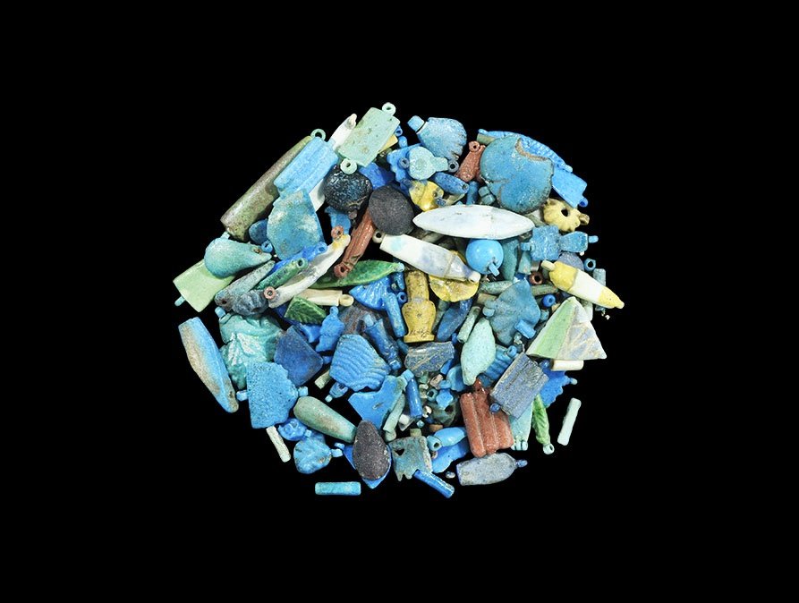 Egyptian Glazed Composition Amulet Group: Amarna Period, 1353-1336 BC. A mixed group of amuletic beads and pendants including fruit, rosettes, rectangular plaques and discoid beads. 44 grams total, 2-37mm (1/10 - 1 1/2"). From a private colle