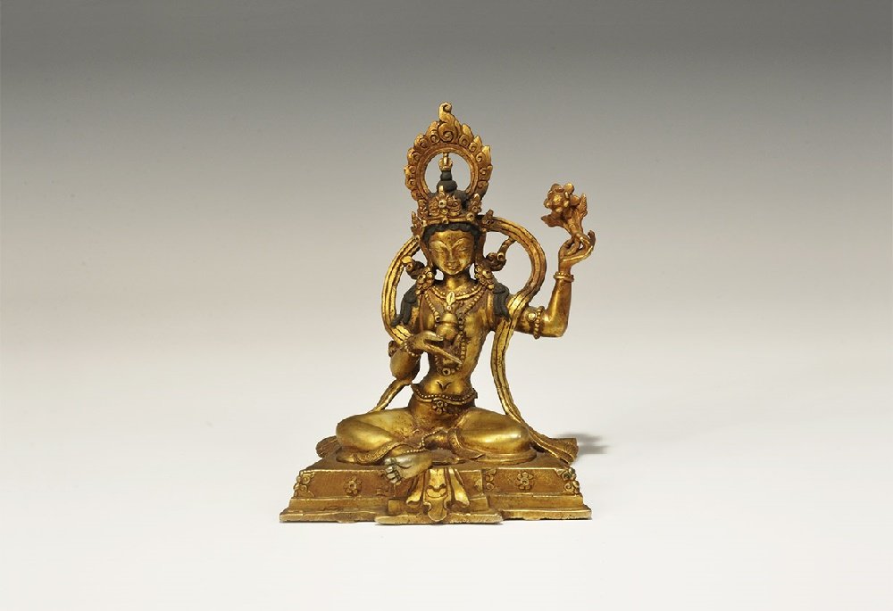Thailand Gilt-Bronze Arya Tara Figurine: 20th century AD. A hollow-cast figurine of Arya Tara sitting on a trapezoidal base with crown of flames and symbols in her hands. 637 grams, 15cm (6"). Property of a London gentleman; acquired 1990-20