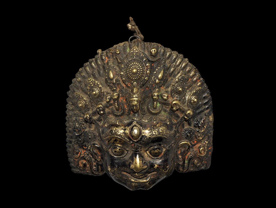 Indian Bronze Bhairava Head Aplique: Nepal, 19th-early 20th century AD. Cast bronze mask of the Hindu god Bhairava shown wearing a crown and with upstanding hair which as various deities and symbols in it; Bhairava has a third eye and fe