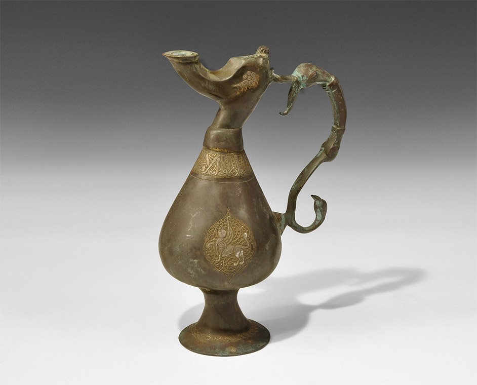Islamic Khorasan Bronze Calligraphic Ewer: 12th century AD or later. A cast vessel with piriform body, flared base, scooped spout, S-curved handle with beast-head at the upper end, bird-head at the lower; silver-inlaid guilloche to the base, r