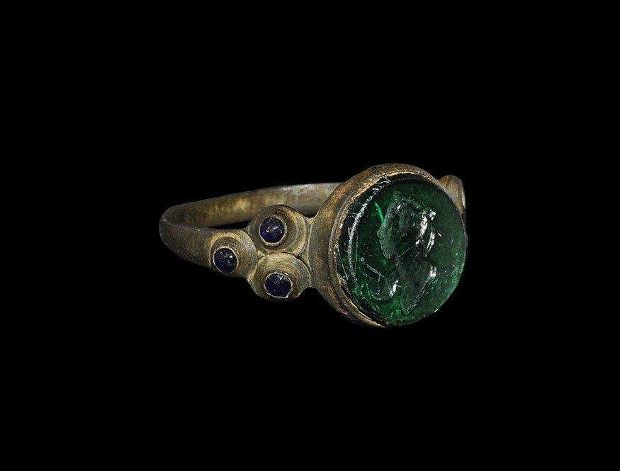 Post Medieval Ring with Green Glass Intaglio: 18th century AD. A narrow D-section bronze hoop, triangular shoulders inset with three blue glass cabochons, discoid cell with inset green glass panel, intaglio motif of a profile female bust with pon