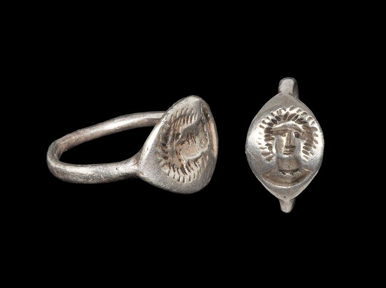 Greek Ring with Face of Apollo: 5th-3rd century BC. A round-section silver hoop supporting a discoid plaque with intaglio mask of Apollo with beams radiating from the head. 2.71 grams, 19mm overall, 16.95mm internal diameter (approx