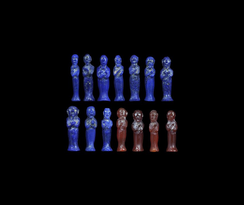 Egyptian Amulet Group: Late Period, 664-332 BC. A mixed group of amuletic standing male figurines, four in carnelian and ten in lapis lazuli, each with an old collector's label to the reverse. 4.34 grams total, 14-16mm (1/2