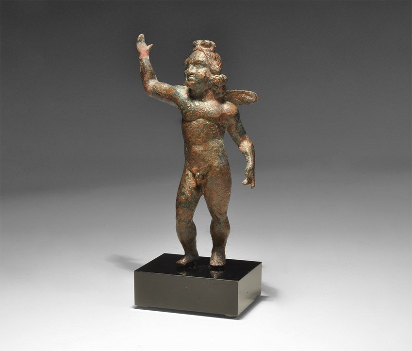 Roman Eros Statuette: 1st century BC-1st century AD. A bronze figure of Eros standing nude with right hand raised, left hand to the side, both modelled open; two small wings to the rear, flowing locks of hair; mounted on a
