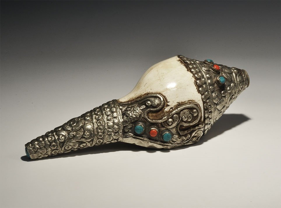 Tibetan Style Mounted Conch Shell: 20th century AD. A large conch shell with silver-coloured metal mounts to the mouth and both ends decorated with a repoussé frieze of vegetation, inset cabochons; hare motif to the mouth; exposed she