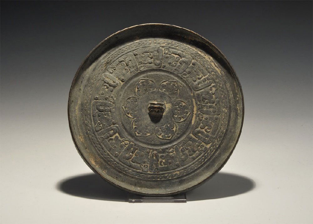 Chinese Style Mirror with Text: An undated archaistic bronze discoid mirror with one polished face, raised rim, central lug, rosette and band of calligraphic text. 784 grams, 17.5cm (6 3/4"). From a central London collection formed
