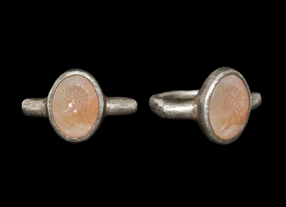 Roman Ring with Sol Invictus Intaglio: 1st-2nd century AD. A round-section silver hoop expanding to keeled shoulders, elliptical cell with inset agate cabochon, intaglio bust of Sol Invictus with radiate crown. 13 grams, 32mm overall, 23.3