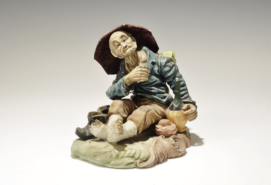 Vintage Spanish Ceramic Figurine: 20th century AD. A hollow figurine of an old man in tattered clothes sitting beneath an umbrella with his boots off, cup in his right hand, loaf and carafe beside his left hip. 915 grams, 18cm (7"). E