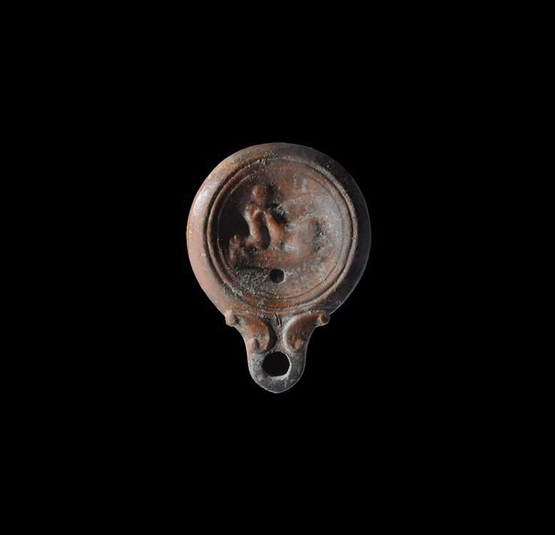 Roman Oil Lamp with Erotic Scene: 2nd century AD. A discoid terracotta lamp with short nozzle and scrolled shoulders, low-relief erotic scene to the discus. From a large oil lamp collection, Surrey, UK; acquired in the 1990s. Cf. Bém