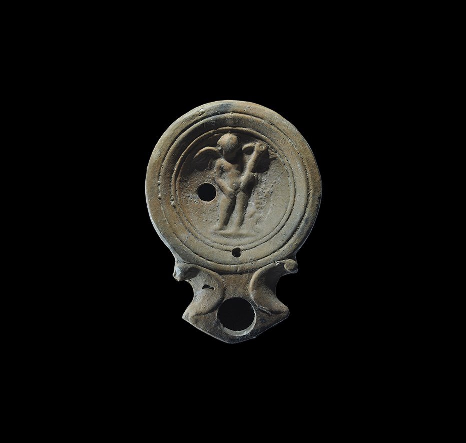 Roman Oil Lamp with Cupid: 1st century AD. A terracotta lamp with deep body, broad nozzle with scrolled shoulders, concentric rings with high-relief figure of Cupid (Eros), winged and with a cockle shell in the left hand. From 