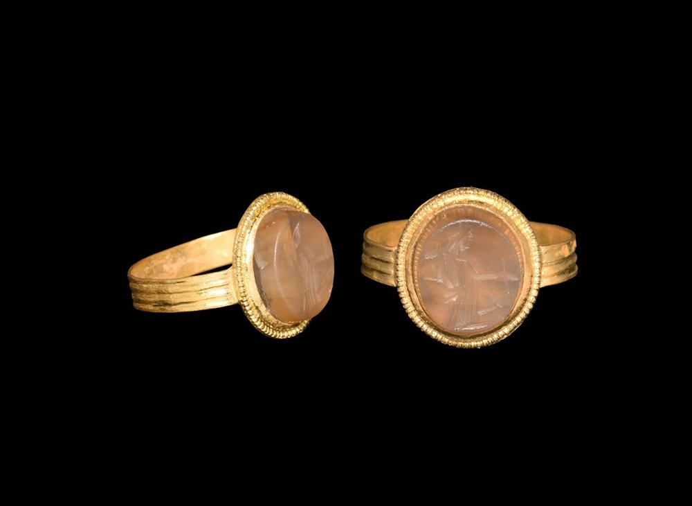 Roman Gold Ring with Fortuna Intaglio: 1st century AD. A flat-section gold hoop with circumferential ribbing, discoid plaque with beaded wire edge, inset banded agate cloison with intaglio image of Fortuna with cornucopia and rudder. Cf. R