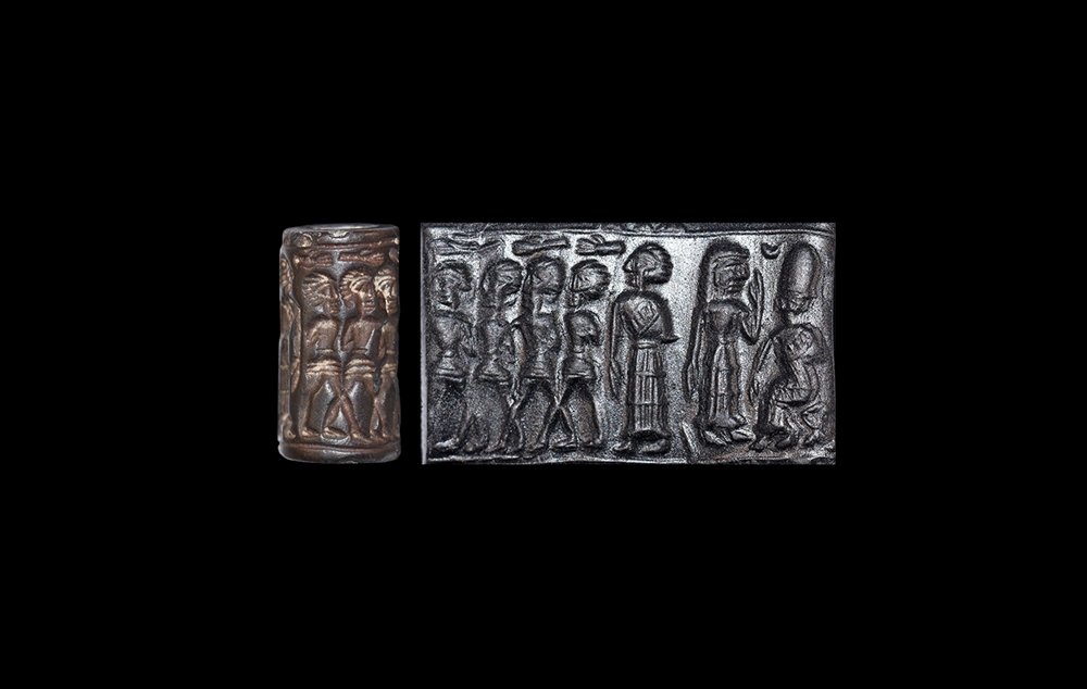 Western Asiatic Old Babylonian Period Cylinder Seal: 18th-17th century BC. A carved haematite cylinder seal with a motif of a central seated deity wearing long dress and very tall hat with attendants gathered behind his throne, a standing female standin