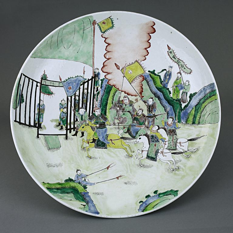 LARGE ANTIQUE CHINESE FAMILLE VERTE CHARGER: Of shallow dish, the interior vibrantly hand painted with a figural scene depicting attendants holding flags and guarding a gate, with armed soldiers on horses fighting each other using spears, swords