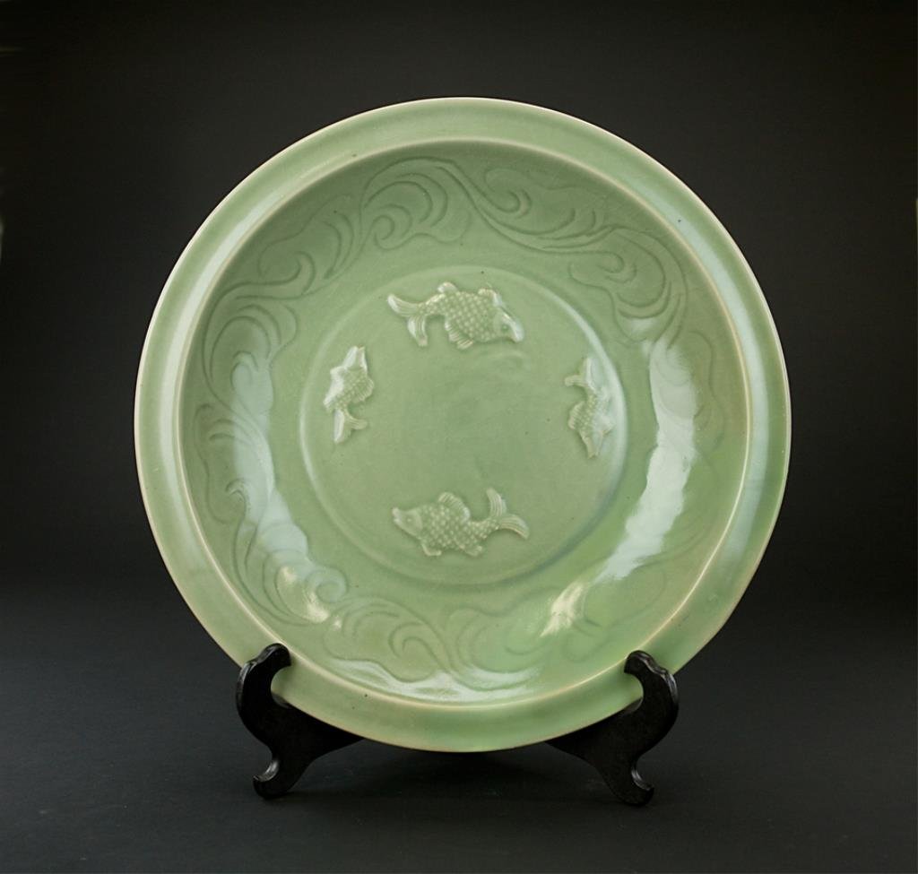 CHINESE CELADON SHALLOW DISH, WITH FOUR FISHES: The center roundel with four raised fish. The interior rim incised with large wave design. The exterior rim decorated with vertical lines. On a wooden stand. Diameter: 16 in.