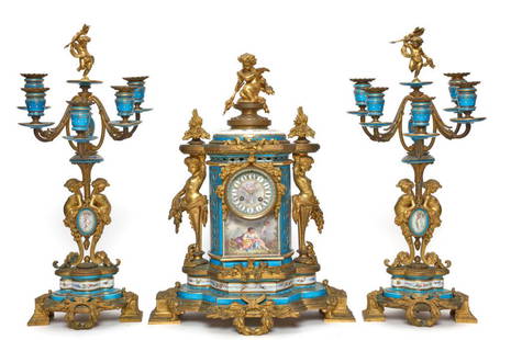 SEVRES ORMOLU THREE-PIECE CLOCK GARNITURE: The mantel clock is surmounted by a cherub with flanking flower finials above columnar bleu celeste clock with painted dial and putto herm caryatids, acanthus and olive leaf base; together with two fo