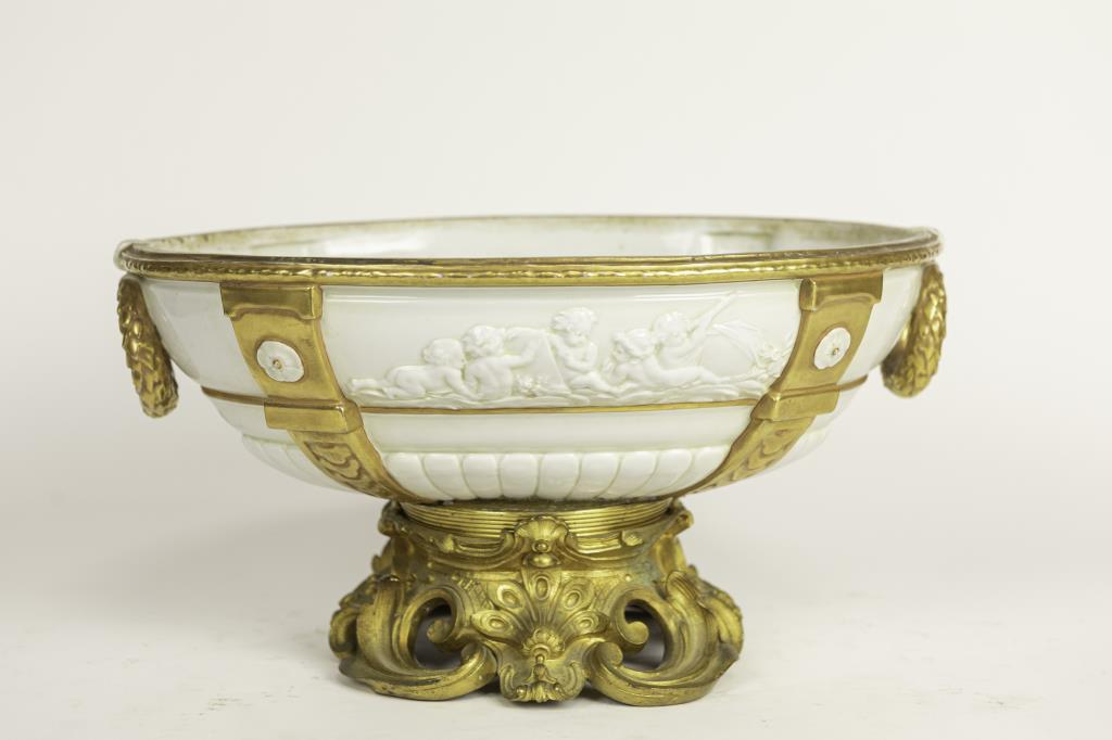 FRENCH WHITE PORCELAIN CENTERPIECE: Beautifully painted with gilt details with cherubim in relief on the body, and on its bottom. Mounted on a gilt bronze base with rococo design, and wide gilt vertical bands, ring handles and rim frame