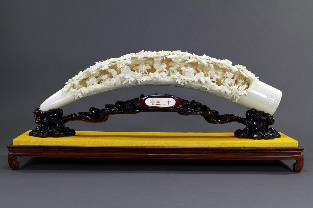 CHINESE CARVING ON STAND, OF THE 12 ZODIAC ANIMALS: L: 22 in. (56cm), simulating an arched tusk, the surface deeply undercut with a dynamic representation of the 12 Chinese zodiac animals beneath pine and prunus trees, on a fitted carved stand with red