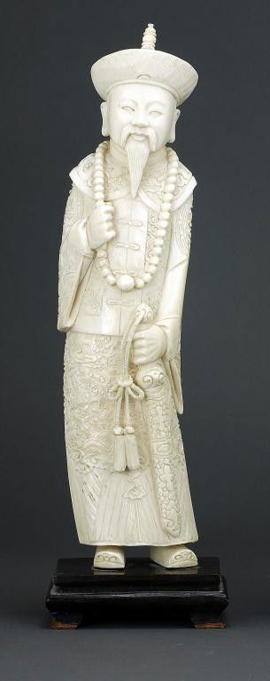 CHINESE CARVED IVORY OF AN EMPEROR: dressed in elaborate court robes and a bonnet, grasping his court necklace in one hand, the other resting on his sword on a wooden stand. H: 10 1/2 in. (26.7cm)