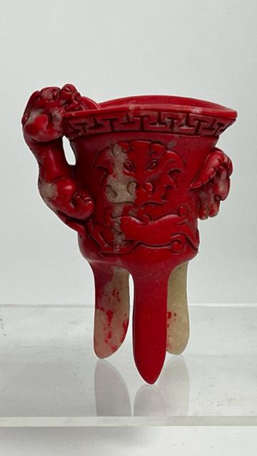AN UNUSUAL CHINESE CINNABAR-COLORED WINE VESSEL: A mythical beast lurks around the cup and lid to form as its handle, the lid with a border of scrolls, body carved, and standing on three tall and slightly flared spear legs.
