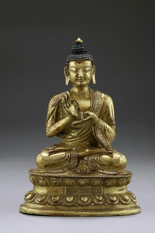 CHINESE GILT-BRONZE FIGURE OF A SITTING BUDDHA: Seated in vajraparyankasana, Both of his hands in mudra. Wearing a pleated robed draped over the left shoulder and falling over the crossed legs. The hair arranged in rows of small pointed whorls and