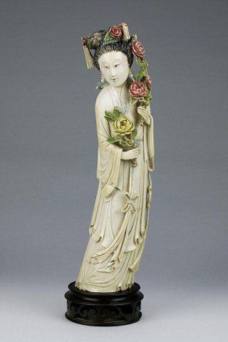 CHINESE POLYCHROMED IVORY FIGURE OF A BEAUTY: Beautifully detailed rendition of an ivory beauty; standing in a modest billowing robe while holding aloft a bloom in her right hand, and a colorful blooming stem in left to match her elaborate floral