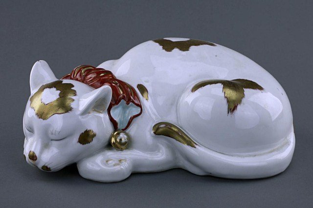 JAPANESE KUTANI FIGURE OF A SLEEPING CAT: 19th century, whimsically hand-painted with a red bow with bell and gold patches. L: 13 1/2 in.