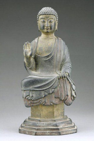 ANTIQUE CHINESE BRONZE OF A SEATED BUDDHA: Cast seated on a raised plinth, with snail-curled hair and a face in a contemplative expression flanked by a pair of long lobes, dressed in a long billowy robe with the left hand resting on its lap wh