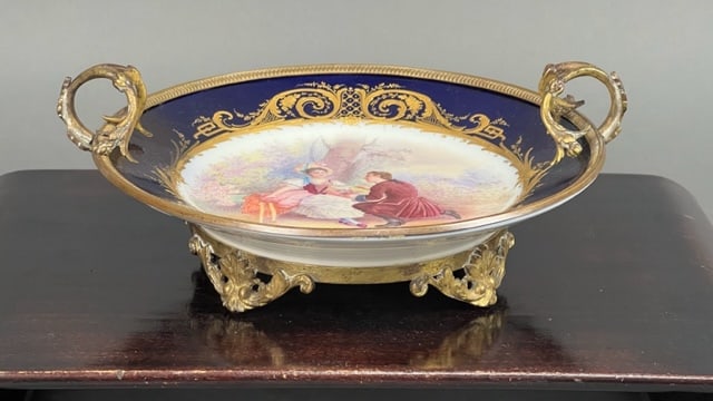 ANTIQUE FRENCH SEVRES COBALT BLUE CENTERPIECE: Of 19th century. Beautifully hand-painted centrally with courting couple, encircled by a gilded border. Gilt bronze mounted with two handles and stamps on the bottom. H: 4 1/2 in. D: 10 3/4 in.
