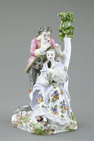 PORCELAIN FIGURE OF LOVERS BY A TREE: The gentleman in the act of placing a flower on the lady's hair as she looks at herself in her hand mirror. Blue crossed marks under the glaze. H: 8 3/4 in.