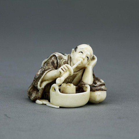 ANTIQUE JAPANESE CARVED IVORY NETSUKE: From the Private collection of Mr. Paul Siegal. Figure of a kneeling man smoking a pipe with a bun and tea pot in front of him. H: 1 in.(2.5cm)