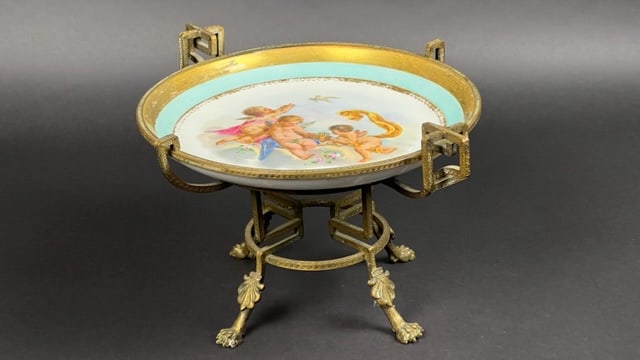 ANTIQUE GILDED & HANDPAINTED PORCELAIN CENTERPIECE: Hand-painted in the center with cherubs, encircled by a gilt rim. Mounted on a gilt bronze stand. Mark on the bottom. H: 8 1/2 in. D: 12 in.