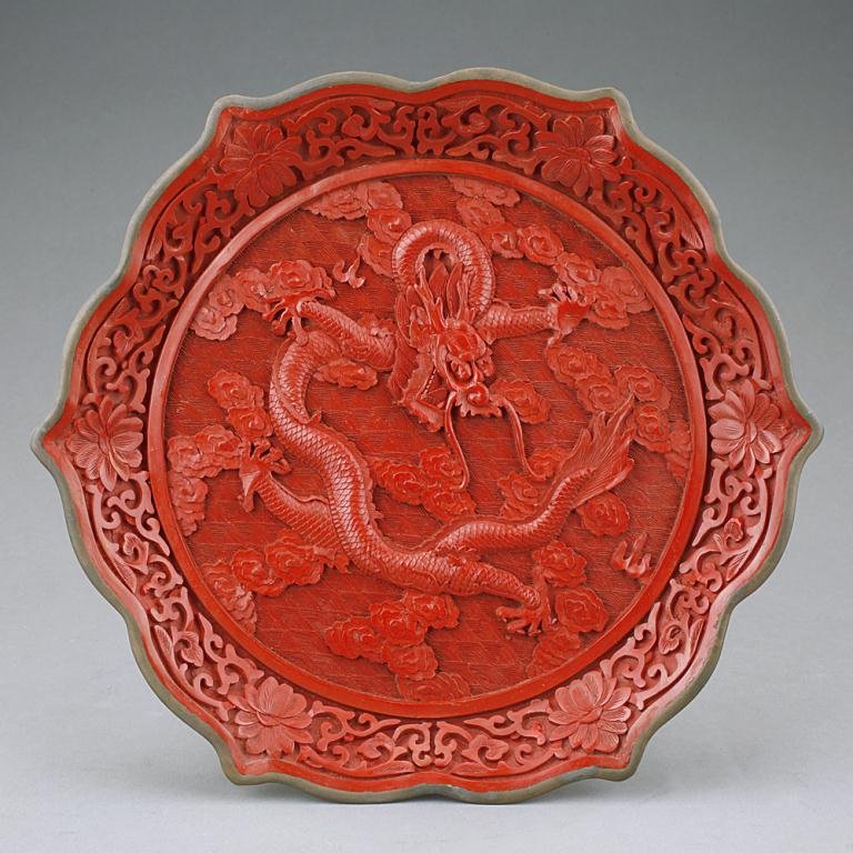 ANTIQUE CHINESE CINNABAR LACQUERED DISH: The medallion at the interior bottom is depicted with a dragon and clouds in relief in a ground of wave pattern while the petal shaped rim is decorated with interlinked flowers. Diameter: 9 in.