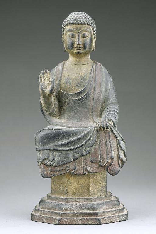 ANTIQUE CHINESE BRONZE OF A SEATED BUDDHA: Cast seated on a raised plinth, with snail-curled hair and a face in a contemplative expression flanked by a pair of long lobes, dressed in a long billowy robe with the left hand resting on its lap wh