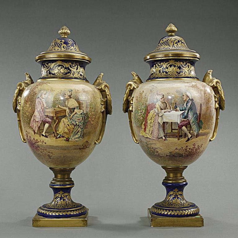 PAIR OF SEVRES COVERED URNS: Pair of 'Sevres-like' covered urns with shouldered ovoid body and gilt on cobalt blue ground. Hand painted design of a couple dining at the table, signed Lucot. The sides with gilt handles and raised