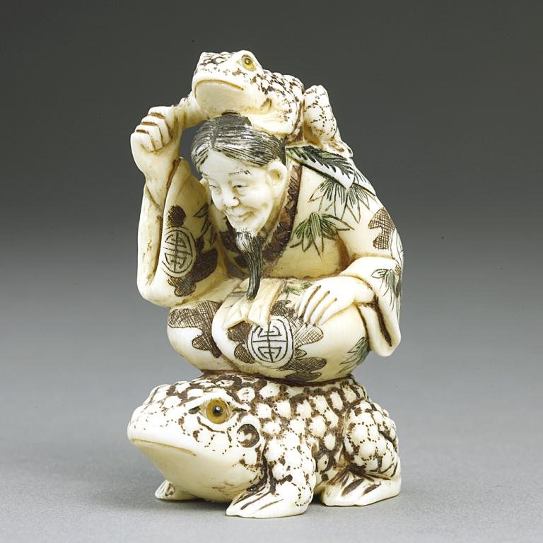 ANTIQUE JAPANESE CARVED IVORY NETSUKE: Of an old man with a long beard and wearing a robe. He is accompanied by two frogs, the smaller rides on the back of his shoulders as he holds on its front leg, while he sits cross legged and rides on