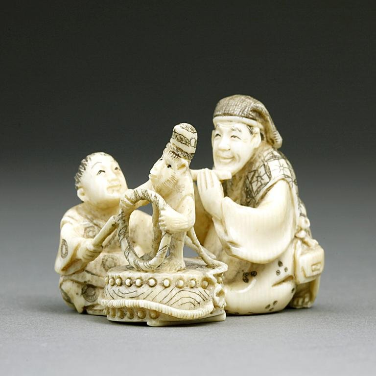 ANTIQUE JAPANESE CARVED IVORY NETSUKE: Of street performers, the man playing his flute while a young girl plays the drum where a monkey is standing on top of it, holding a ring. signature in red, framed at the base. H: 1 1/4 in.