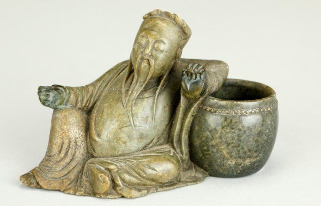 CARVED GREEN SOAPSTONE FIGURE OF A MAN: In a reclined position and leaning against a large vase. With a long wispy beard, he has a serene expression on his face. Stone is of a fine deep olive green color. H: 3 in., L: 4 3/4 in.