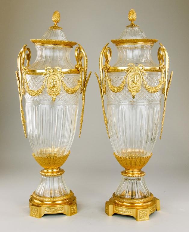 PAIR OF LARGE BACCARAT-STYLE LIDDED URNS: Comprising of cut glass and gilt bronze. Raised upon a gilt bronze four-footed base with cast leaf design rising up from a band around the top of the foot; gilt bronze band around the shoulder with fl